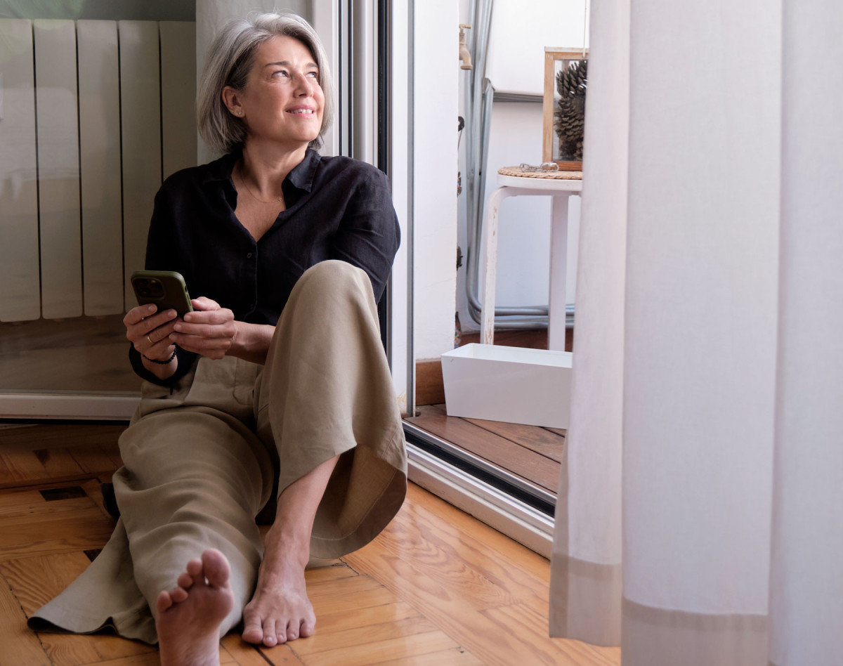 Frau um die 60 sitzt gesund, gelassen, barfuß auf einem Holzboden. Lächelnd, mit Handy in der Hand.